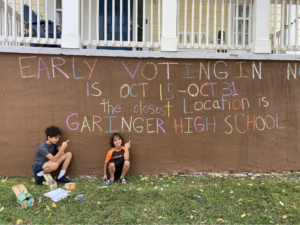 Two kids sitting in front of their chalk drawing promoting early voting.